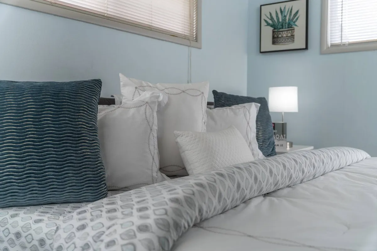 A serene bedroom scene featuring a comfortable bed dressed with decorative pillows and soft linens, accompanied by a glowing table lamp and subtle wall art. Soft natural light filters in, creating a warm and inviting atmosphere, perfect for unwinding after a day of exploration.