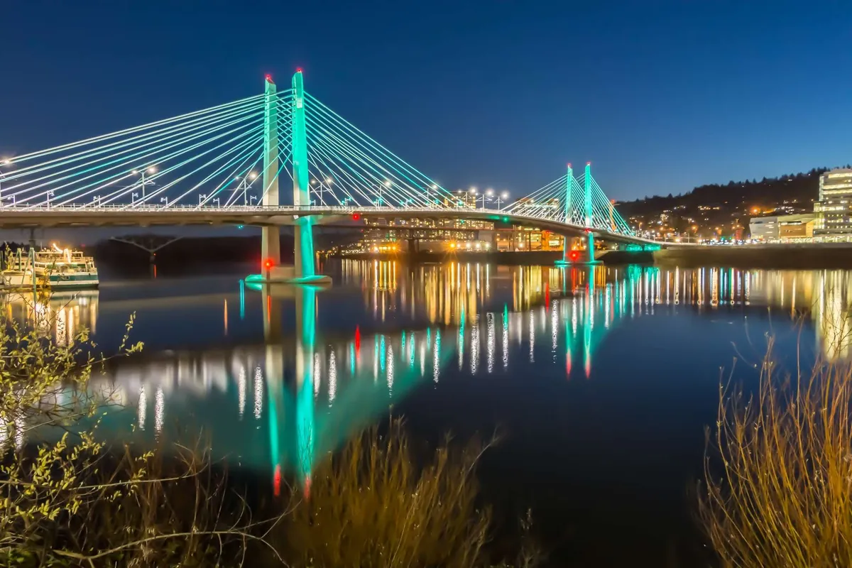 A beautiful nighttime exterior view of a suspension bridge reflected in calm water, with city lights twinkling in the background. The scene is serene and picturesque, inviting thoughts of evening strolls and outdoor adventures near the waterfront.