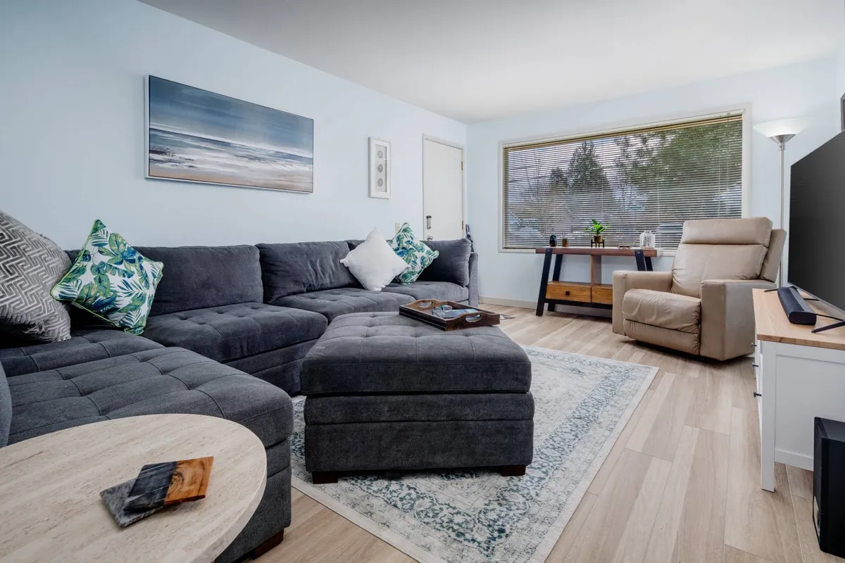 The image showcases a bright and inviting living room featuring a plush sectional sofa adorned with colorful accent pillows, a stylish coffee table, modern artwork on the wall, and a large window streaming in natural light. The space feels cozy and perfect for unwinding or socializing.