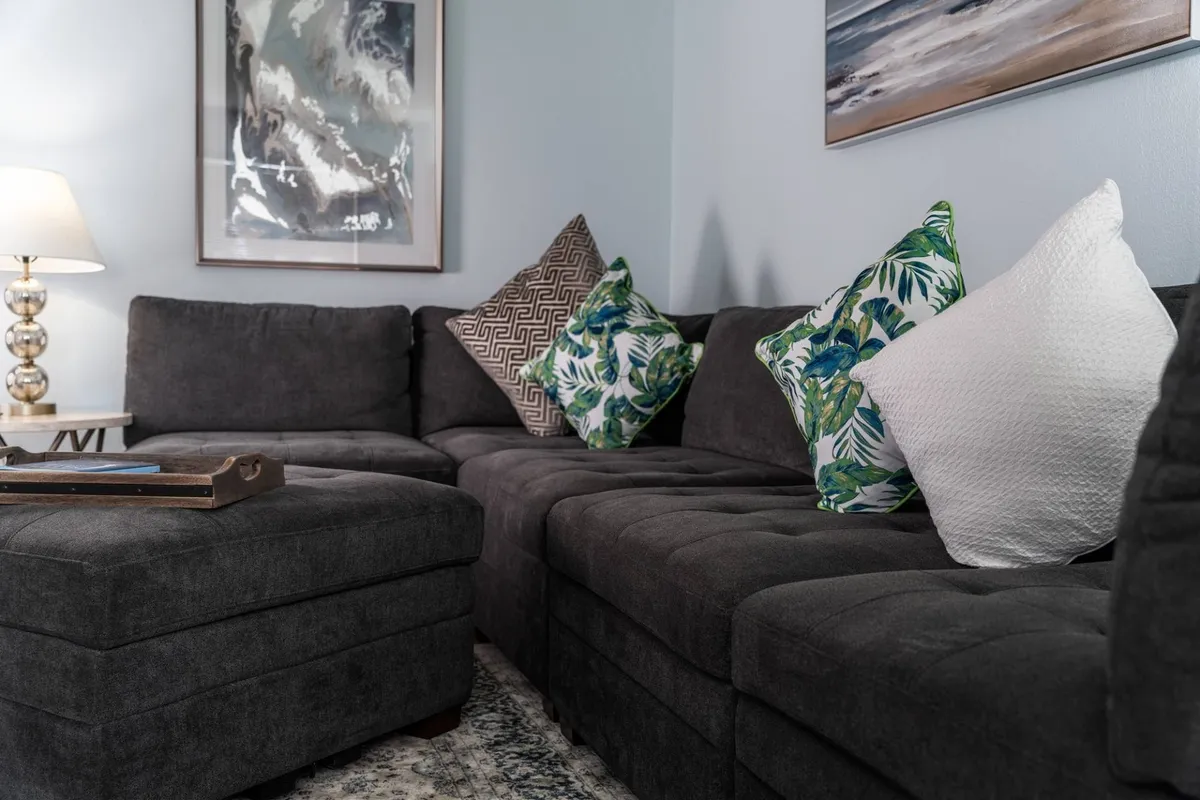 This image features a welcoming sectional sofa accented with decorative pillows, a table lamp that casts a warm glow, and framed artwork on the wall. The style is contemporary yet inviting, with ample natural light streaming into the room, emphasizing the fresh ambiance and comfortable atmosphere. The overall layout hints at an open living area ideal for both relaxation and socializing.