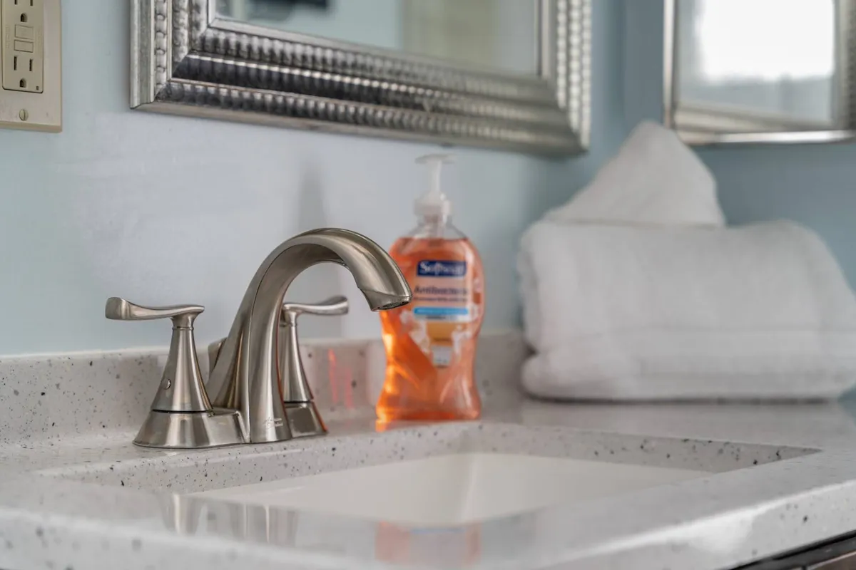 This image features a modern and clean bathroom sink area, with a sleek faucet, soap dispenser, plush towels, and a large mirror. The setting is bright and inviting, evoking a sense of freshness and attention to detail.