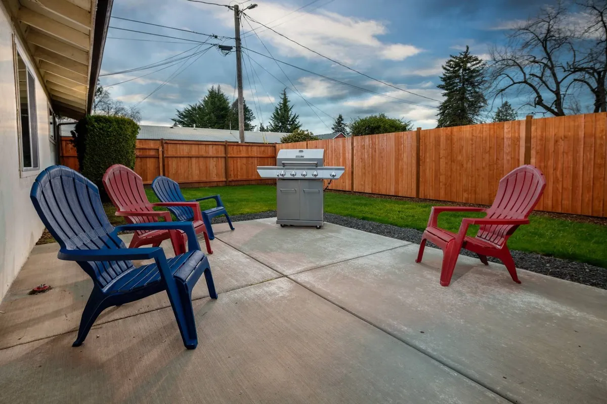 A fenced backyard featuring a BBQ grill and an arrangement of colorful chairs set up for socializing. The space looks inviting for casual gatherings, barbecues, and simply relaxing outdoors. The sunset lighting adds a warm, lively ambiance, making it an ideal spot for al fresco dining or unwinding with family and friends.