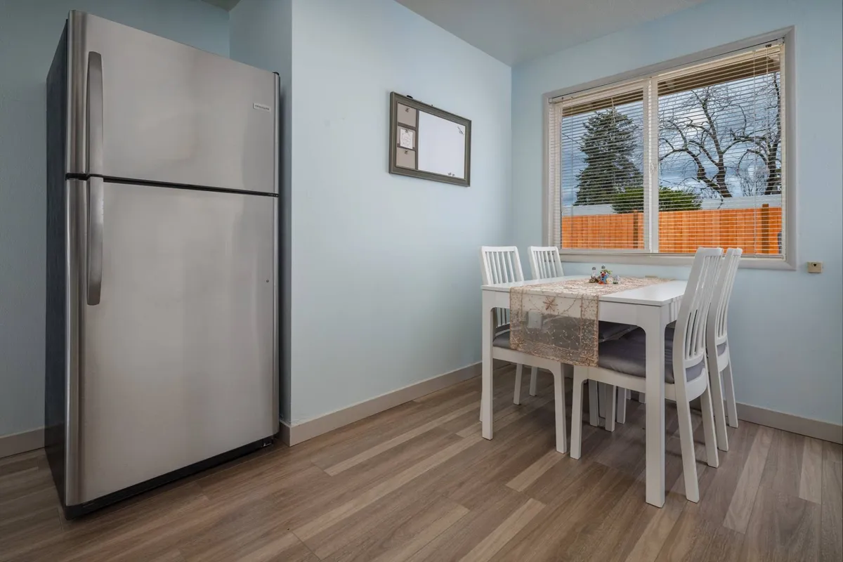 A bright, multi-functional space showcasing a stainless steel refrigerator, a crisp white dining table, soft light blue walls, and a large window that fills the room with sunlight. The setting is cozy and modern, suggesting a seamless transition from entryway to communal living spaces.