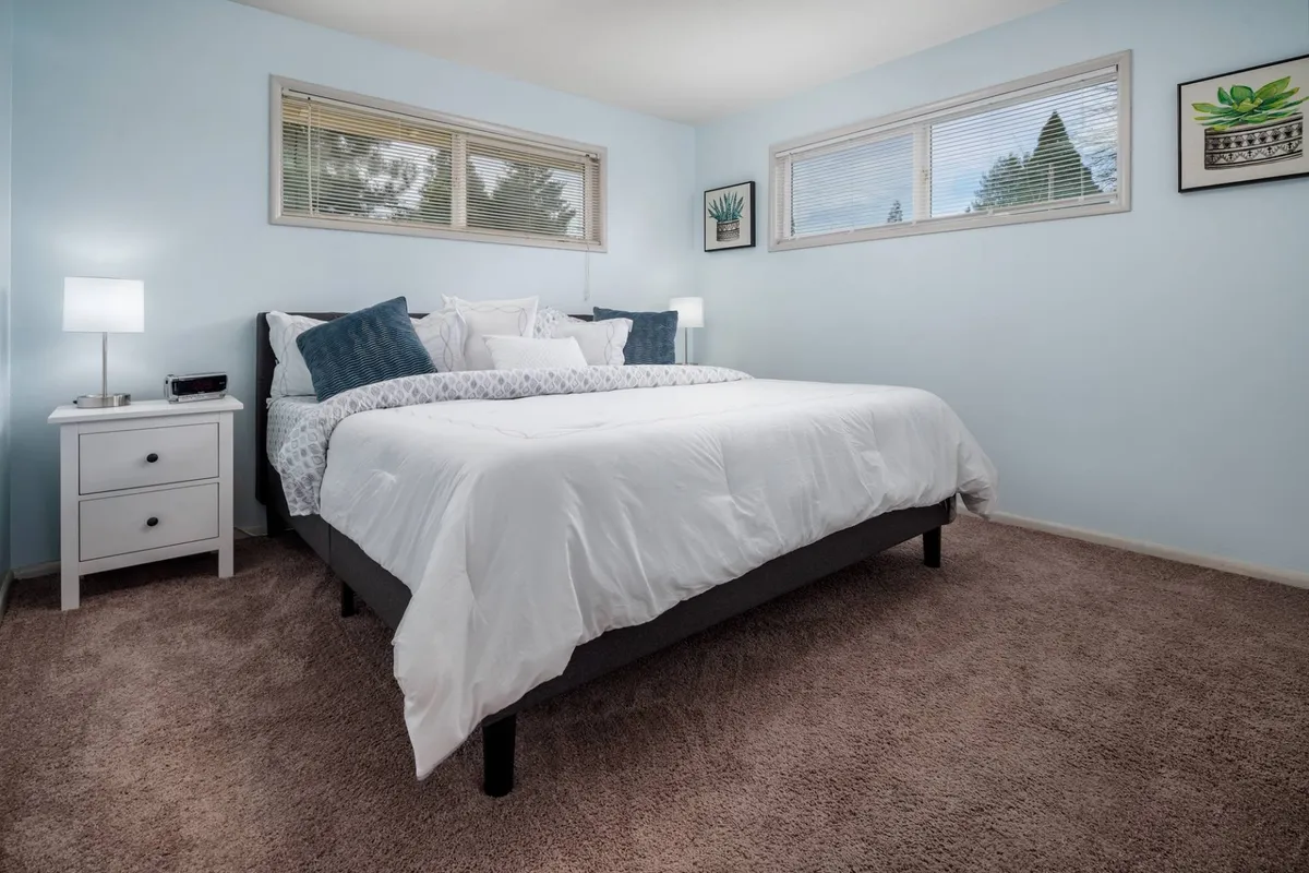 A bright, serene bedroom is shown with a cozy bed, decorative pillows, a nightstand, and large windows allowing natural light to pour in. The overall atmosphere communicates comfort and morning rejuvenation.